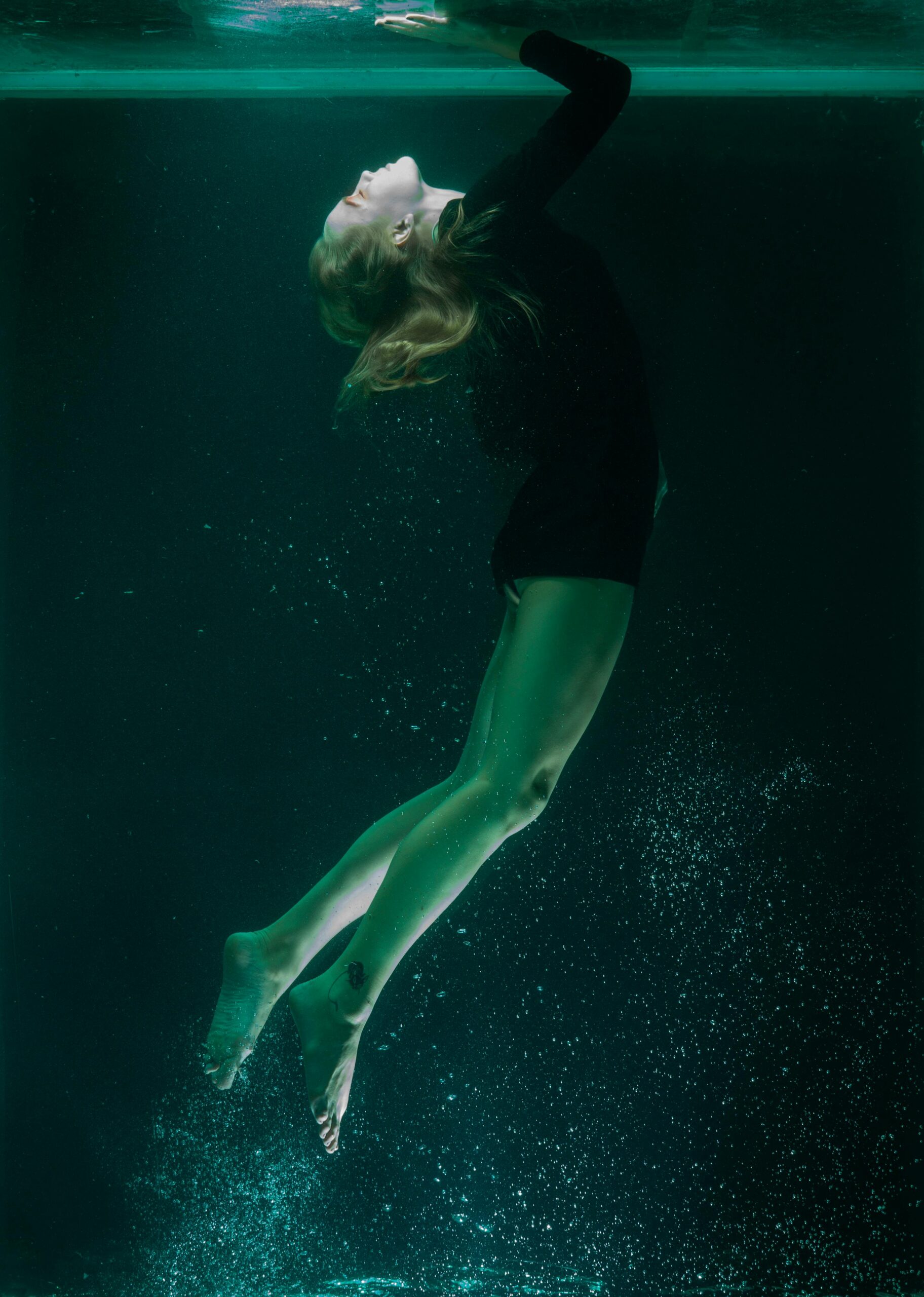 Elegant underwater shot of a woman floating in a serene pose, captured in dim turquoise light.